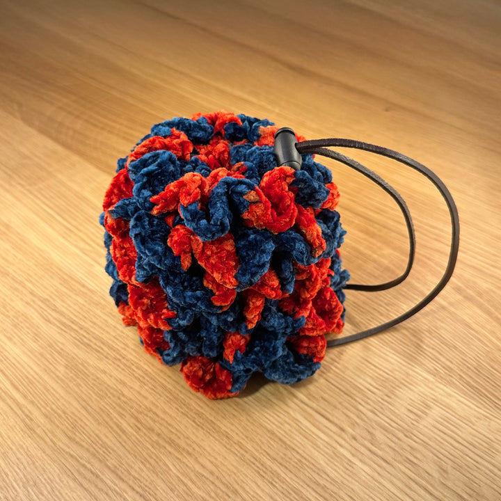 Crochet dragon egg dice bag made from navy and red velvet yarn with chocolate leather drawstring and black plastic toggle, shown closed.