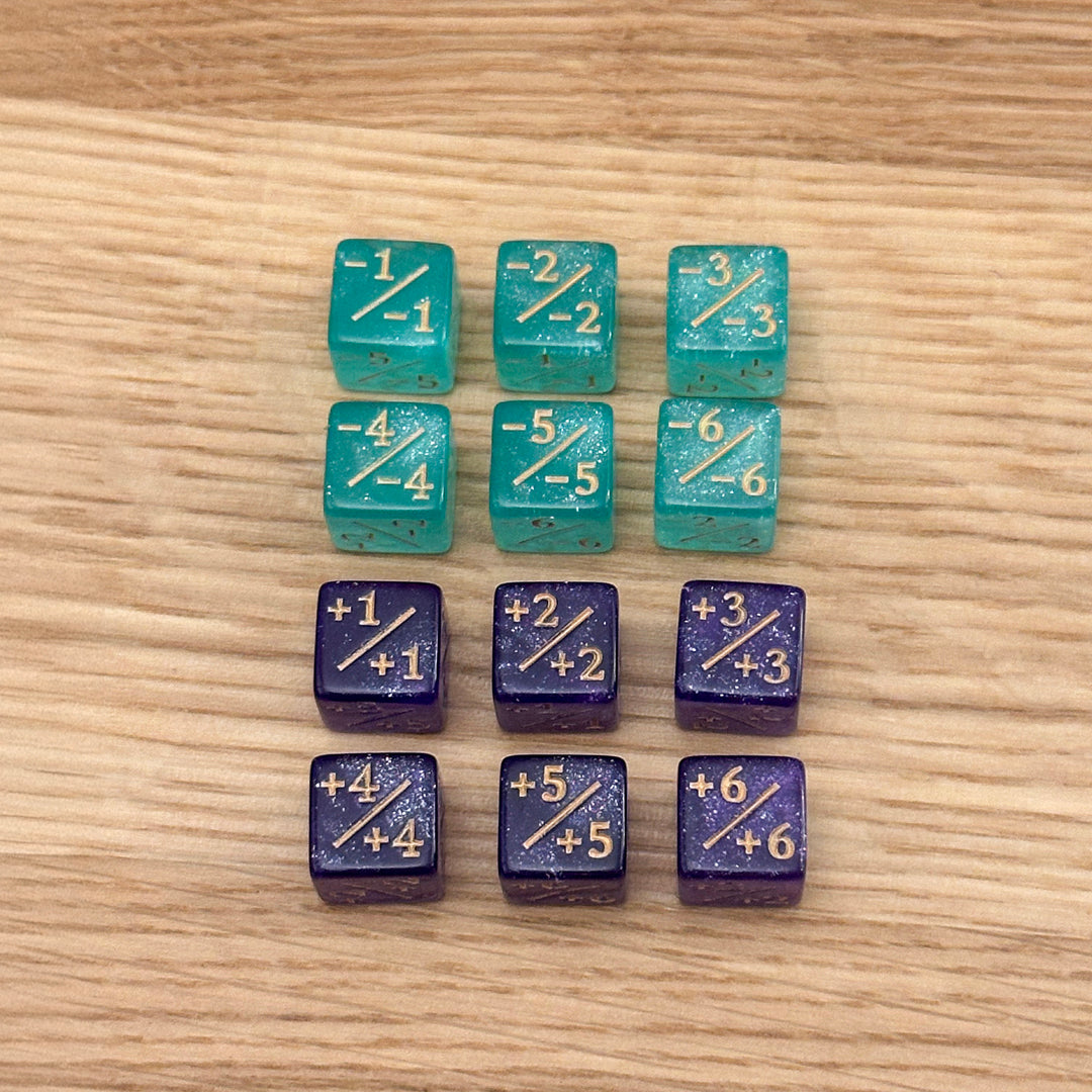 Set of 12, 12mm six-sided counter dice, including six aqua glitter +/+ dice with gold infill and six purple glitter -/- dice with gold infill, used for tracking game modifiers.