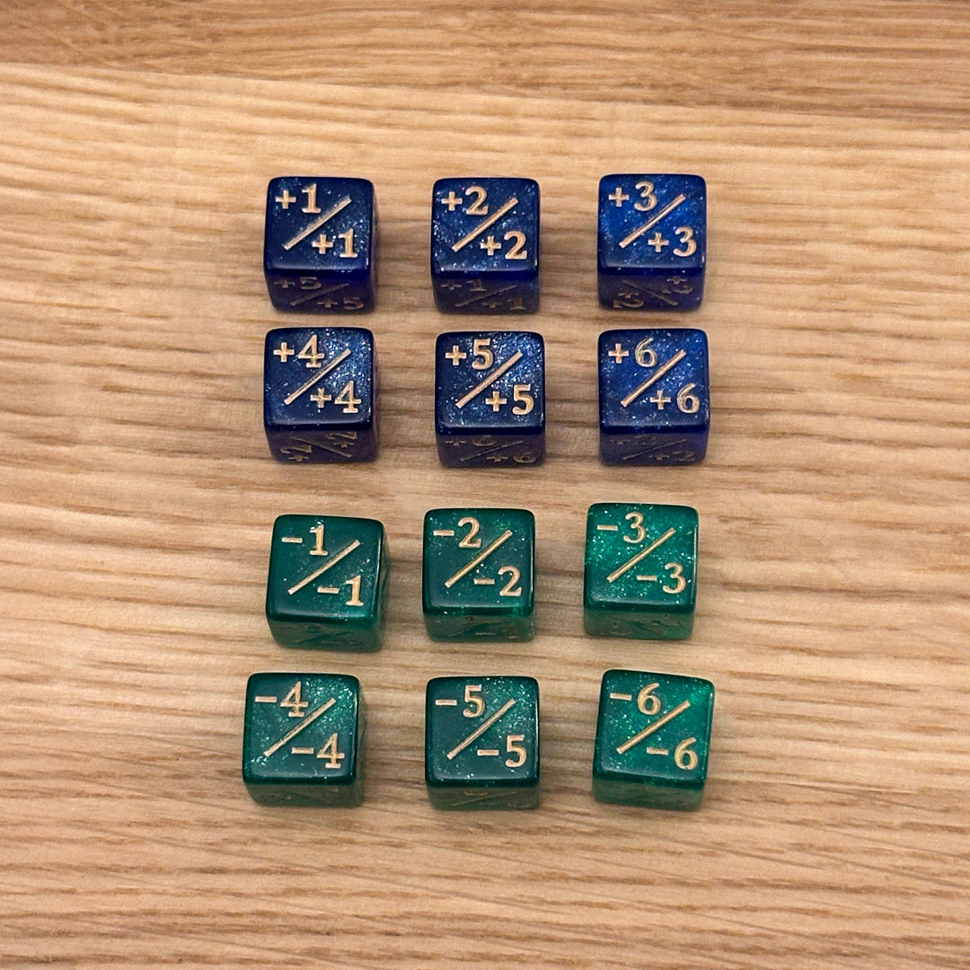 Set of 12, 12mm six-sided counter dice, including six blue glitter +/+ dice with gold infill and six emerald green glitter -/- dice with gold infill, used for tracking game modifiers.