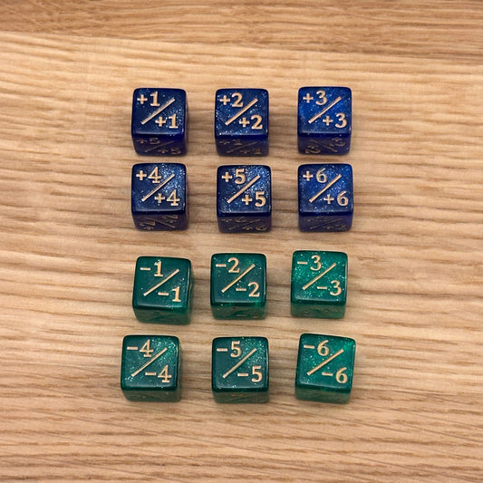 Set of 12, 12mm six-sided counter dice, including six blue glitter +/+ dice with gold infill and six emerald green glitter -/- dice with gold infill, used for tracking game modifiers.