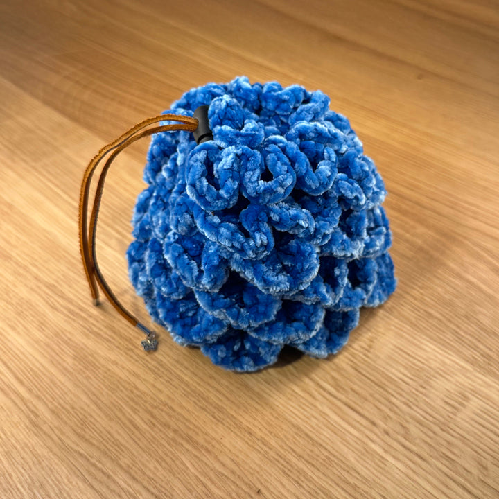 Crochet dragon egg dice bag made from blue velvet yarn with tan leather drawstring and black plastic toggle, shown closed.