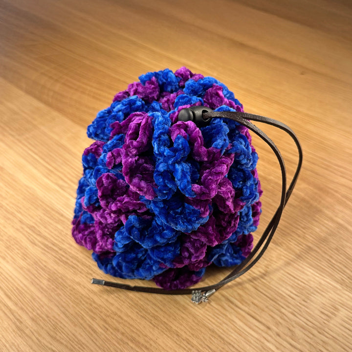 Crochet dragon egg dice bag made from royal blue and purple velvet yarn with chocolate leather drawstring and black plastic toggle, shown closed.