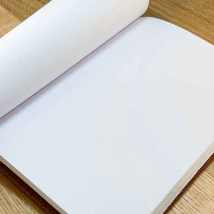 Open hand-stitched leather journal with soft, flexible cover rolled back, revealing blank 80gsm artist-grade white paper.